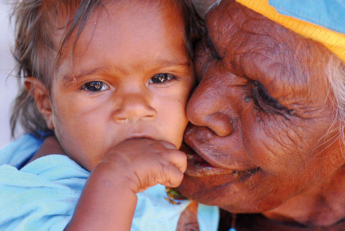 Colour photograph of a close-up shot of an elderly woman affectionately pressing her cheek to a young child's.