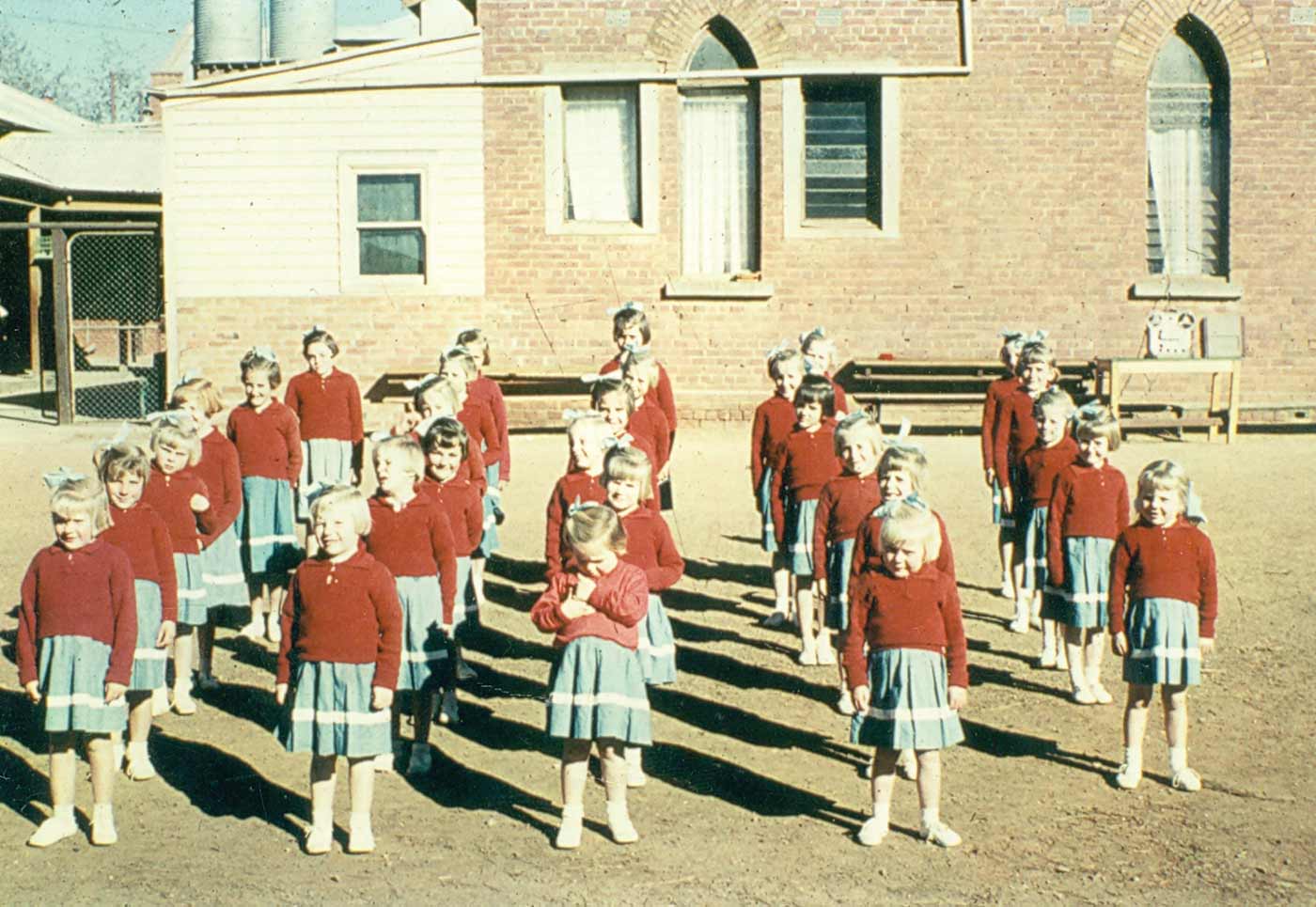Colour photograph showing five rows of young girls lined up outside a brick building. The girls wear red jumpers over blue skirts with a horizontal white line above the knee. Each girl wears white shoes and socks and a blue ribbon in her hair. - click to view larger image