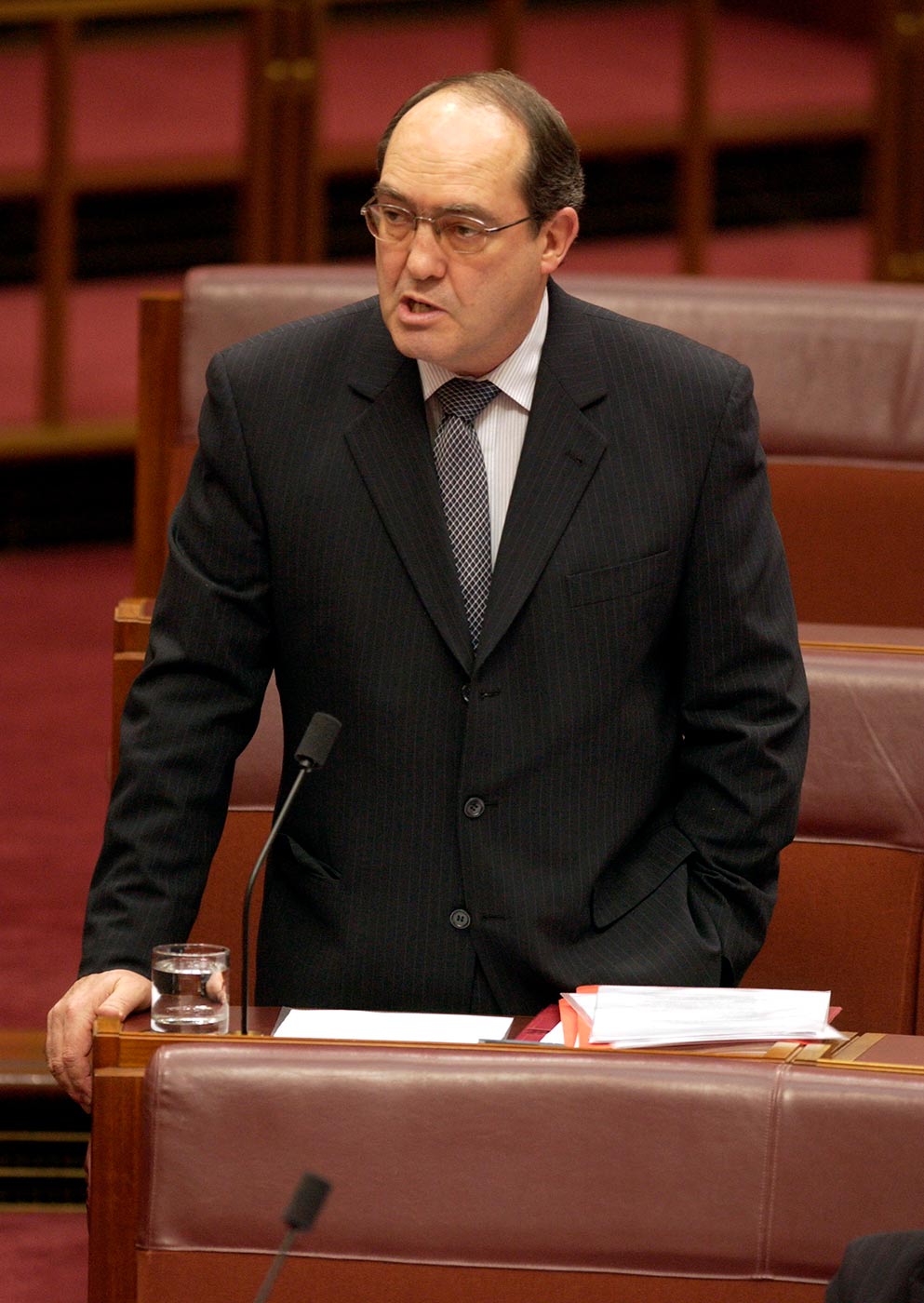 Colour photograph showing a man wearing a suit, leaning on a desk to address a parliamentary chamber. - click to view larger image