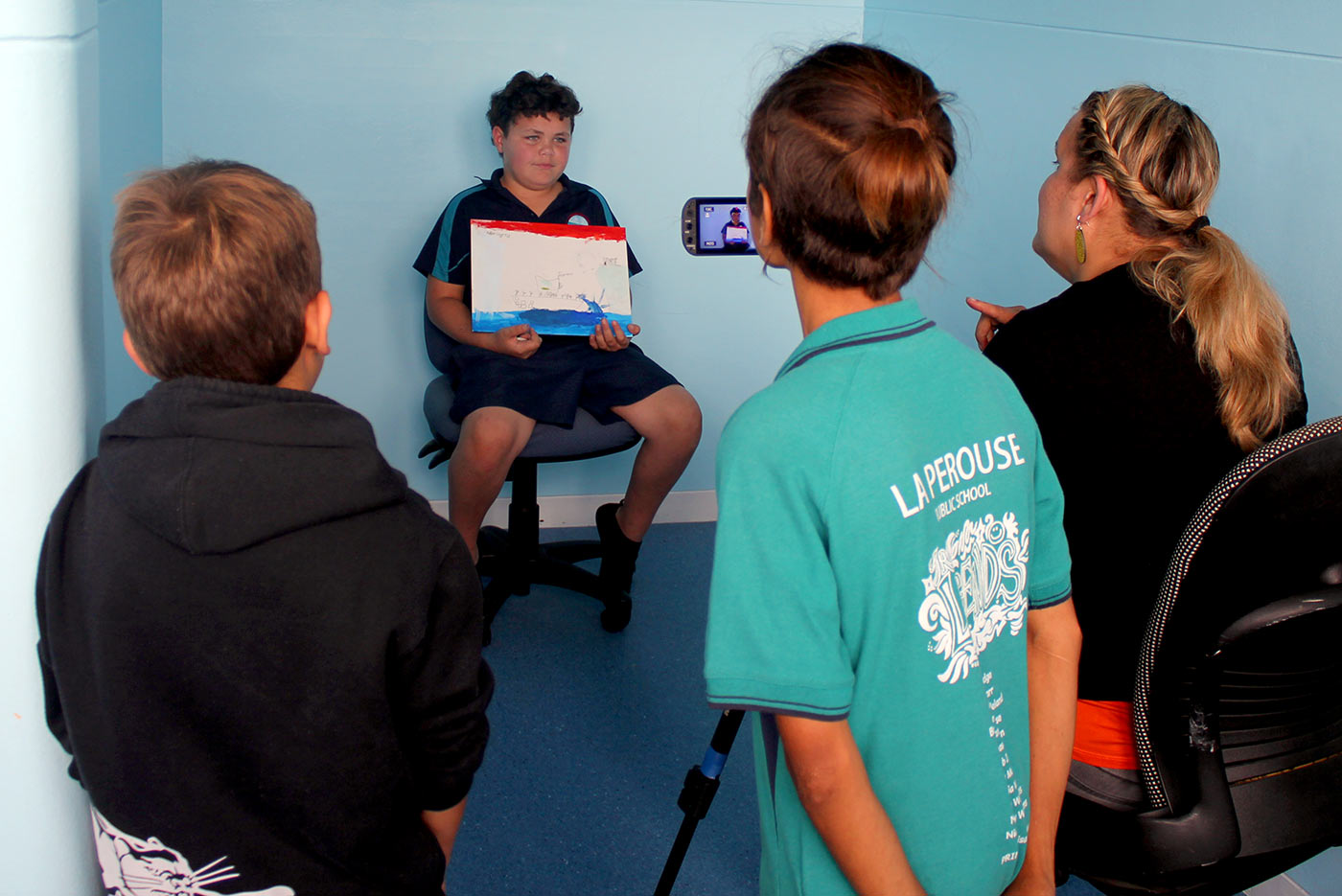 A boy in a school uniform being filmed presenting his painting while two other students and a female interviewer watch on.. - click to view larger image