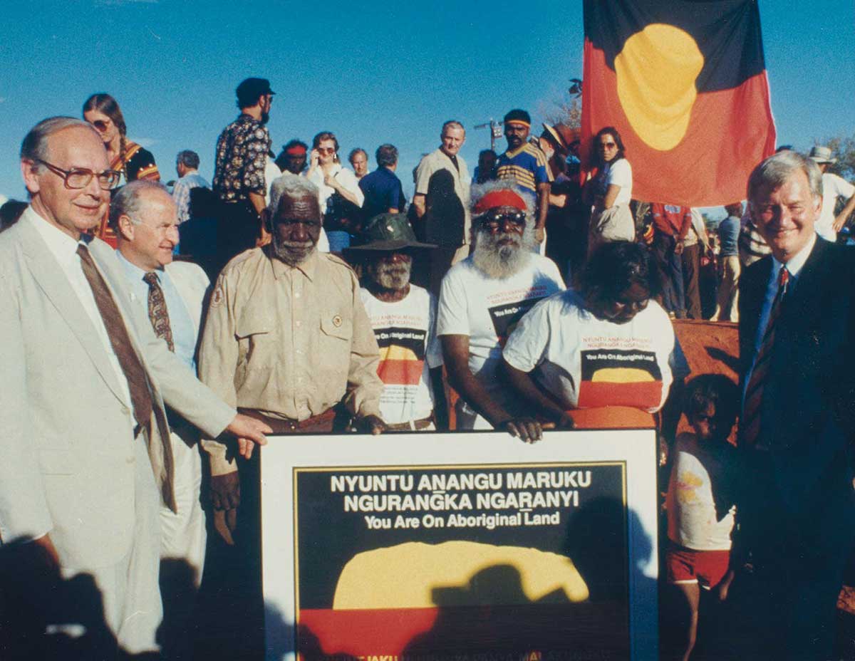 Colour photo of a crowd of people with the Aboriginal Australian flag in the background. In the foreground are several people gathered around a large sign with the words 