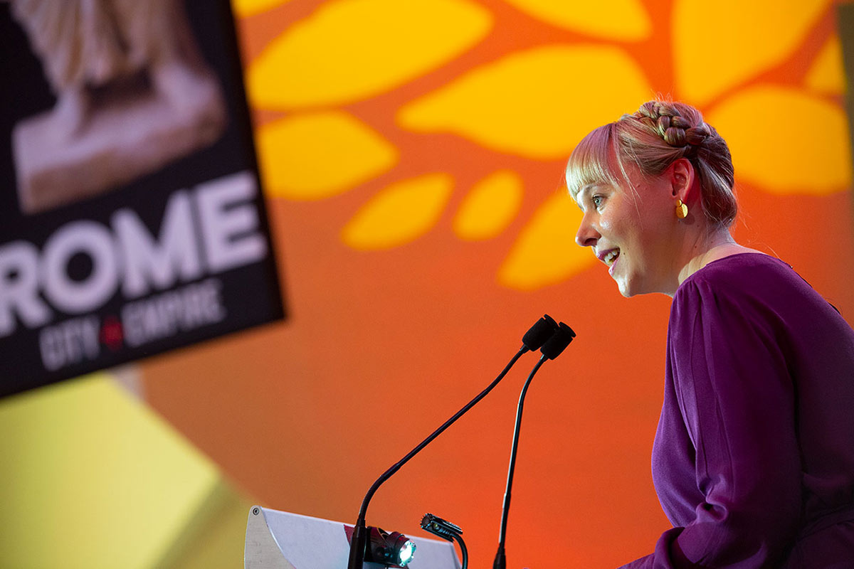 A woman is speaking into a microphone with the 'ROME. CITY+EMPIRE' exhibition banner in the background. - click to view larger image