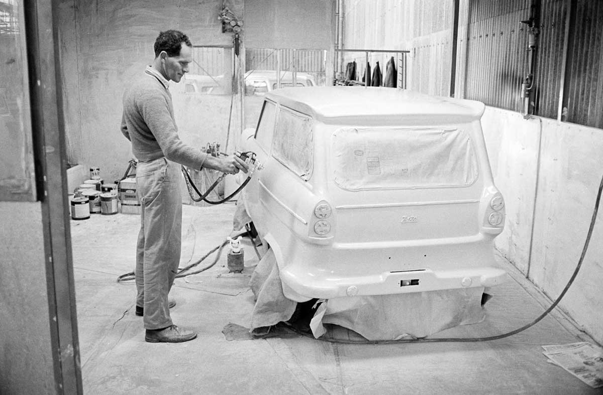 A black and white photo of a worker facing a small white car body. - click to view larger image
