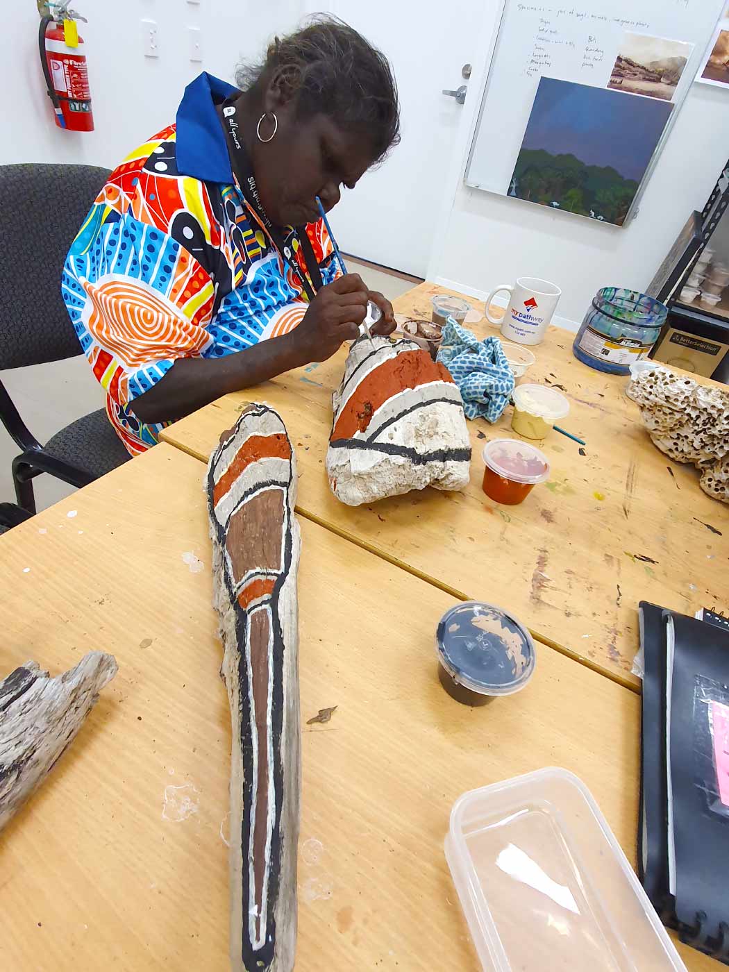 Colour photo of a female artist painting with ochre pigments onto pieces of driftwood in a studio. - click to view larger image