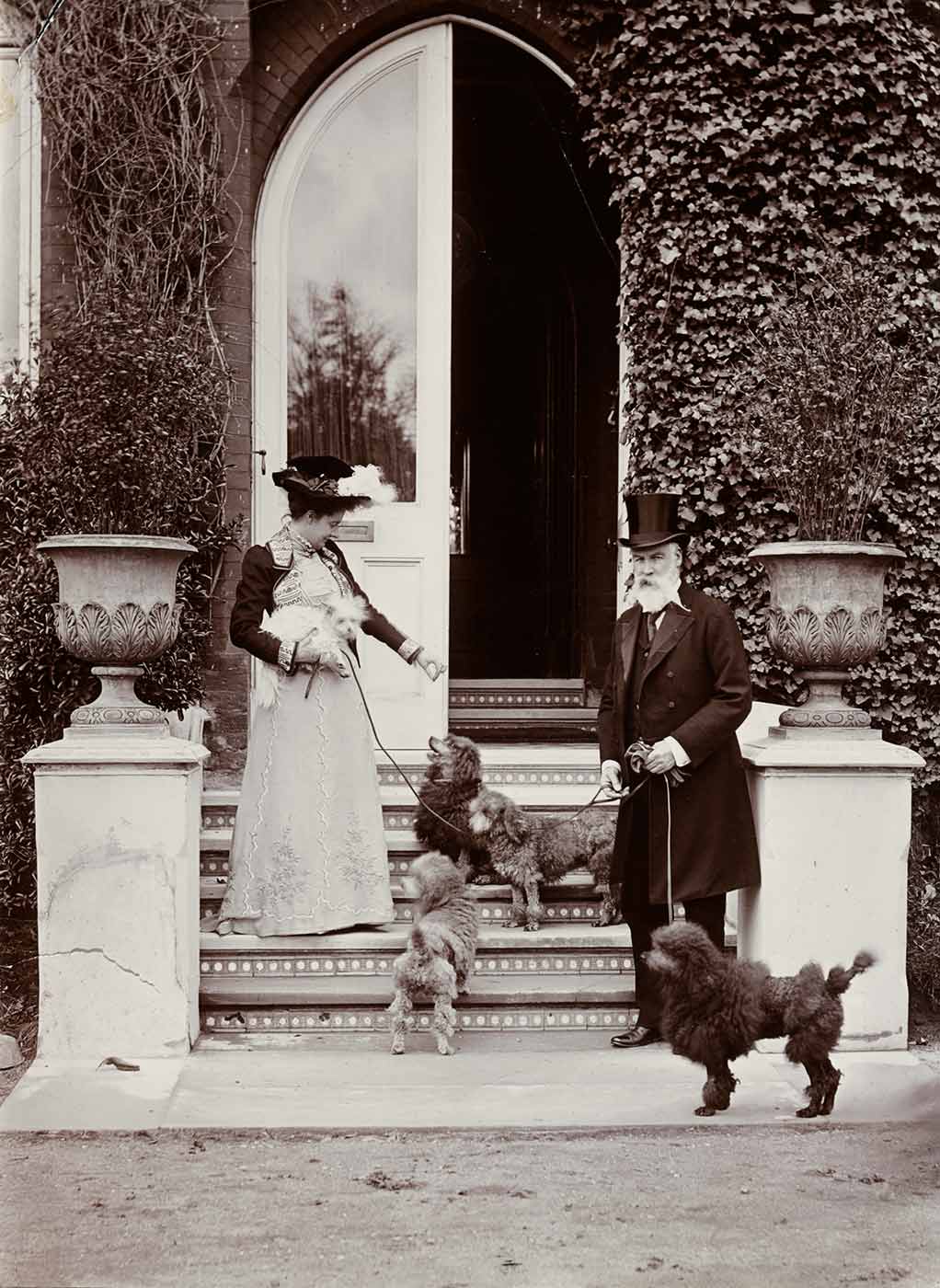 A woman standing on the steps to an entrance wearing a hat and holding one dog with three other dogs on the steps with her and a gentleman wearing a top hat with one dog on a lead standing on the bottom step. - click to view larger image