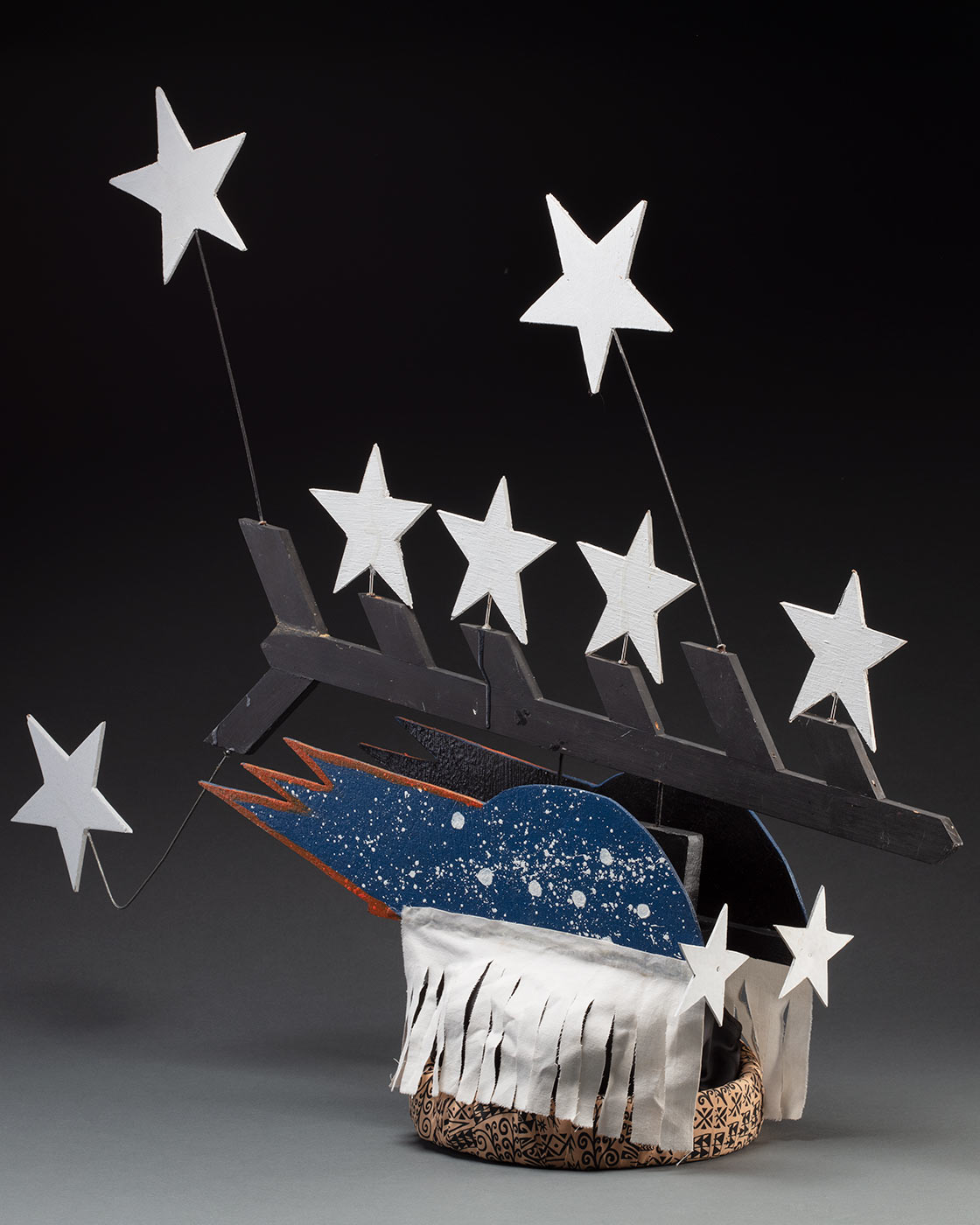 A headdress featuring seven stars protruding from a black painted piece of board forming half a fishbone pattern. It is connected to two pieces of board painted blue with white splatter and seven stars on each side with a white fabric fringe at the bottom. It is all attached to a fabric wrapped headband in the centre.