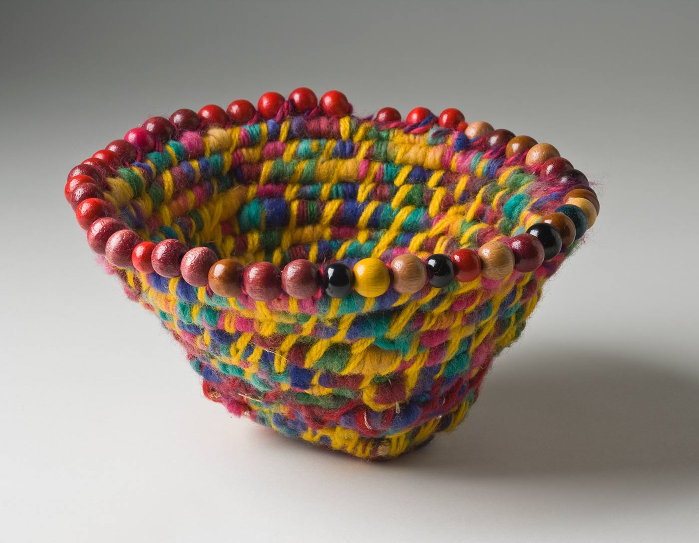 A funnel shaped basket of multicoloured coiled yarn over plant fibre with a beaded edge. The yarn is rainbow coloured with a dominant yellow tone. The maroon yarn used at the base has small silver sequins in it. The round wood beads attached at the top edge of the basket are mostly red and brown with a few green, yellow, black and natural coloured ones. - click to view larger image
