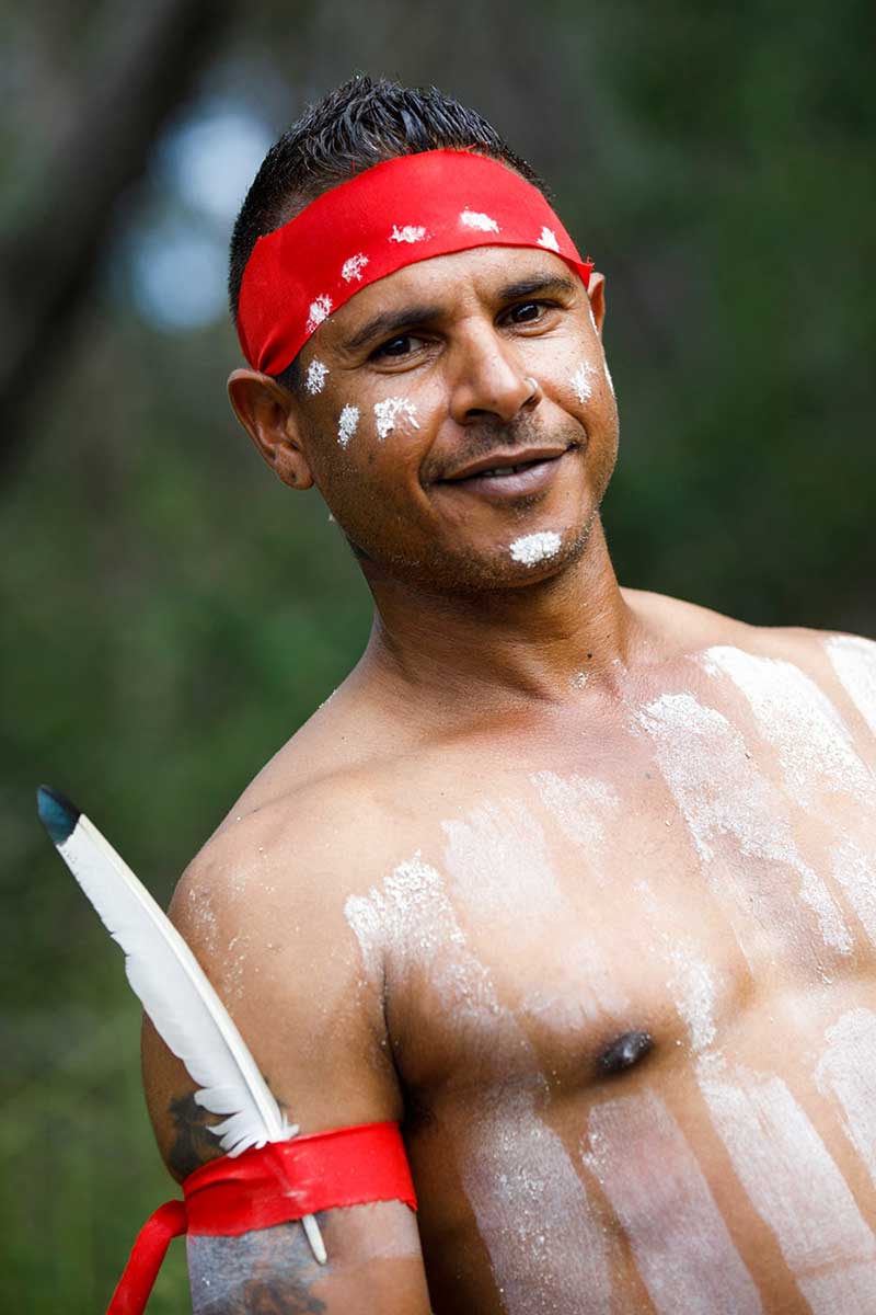Colour photograph of a man smiling and posing for the camera . He is wearing a red bandana and body paint. - click to view larger image