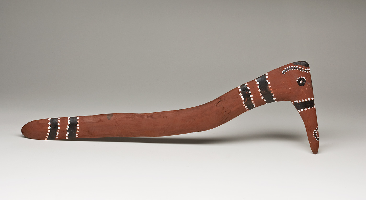 A hooked wooden boomerang with a curved handle, decorated to look like a bird. The boomerang is painted red all over and is plain on the flat side. On the convex side the corner of the hook has a black circle with a white dot inside it plus white dots around part of the edge. The point of the hook has a black stripe and a black curve outlined with white dots, while at the edge of the corner there is a wavy black line edged in white dots and a black semicircle. At the neck and base of the handle are two black stripes edged and divided by lines of white dots. Below the neck of the handle there is a horizontal jagged crack in the wood. - click to view larger image