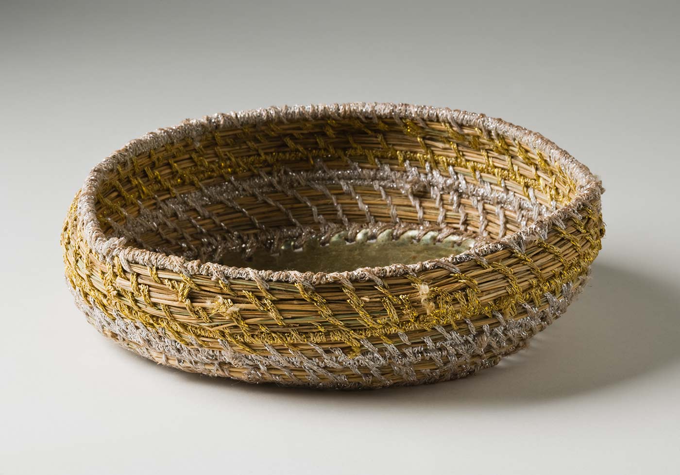 A shallow circular metallic yarn and plant fibre basket with metal base. The base is made of a blue 'Fray Bento / Steak and Kidney Pie' tin lid which has been attached to the yarn section with silver coloured metallic yarn through holes punched in the tin. The basket has silver and gold coloured metallic yarn spaced out so the plant fibre centre can be seen. - click to view larger image