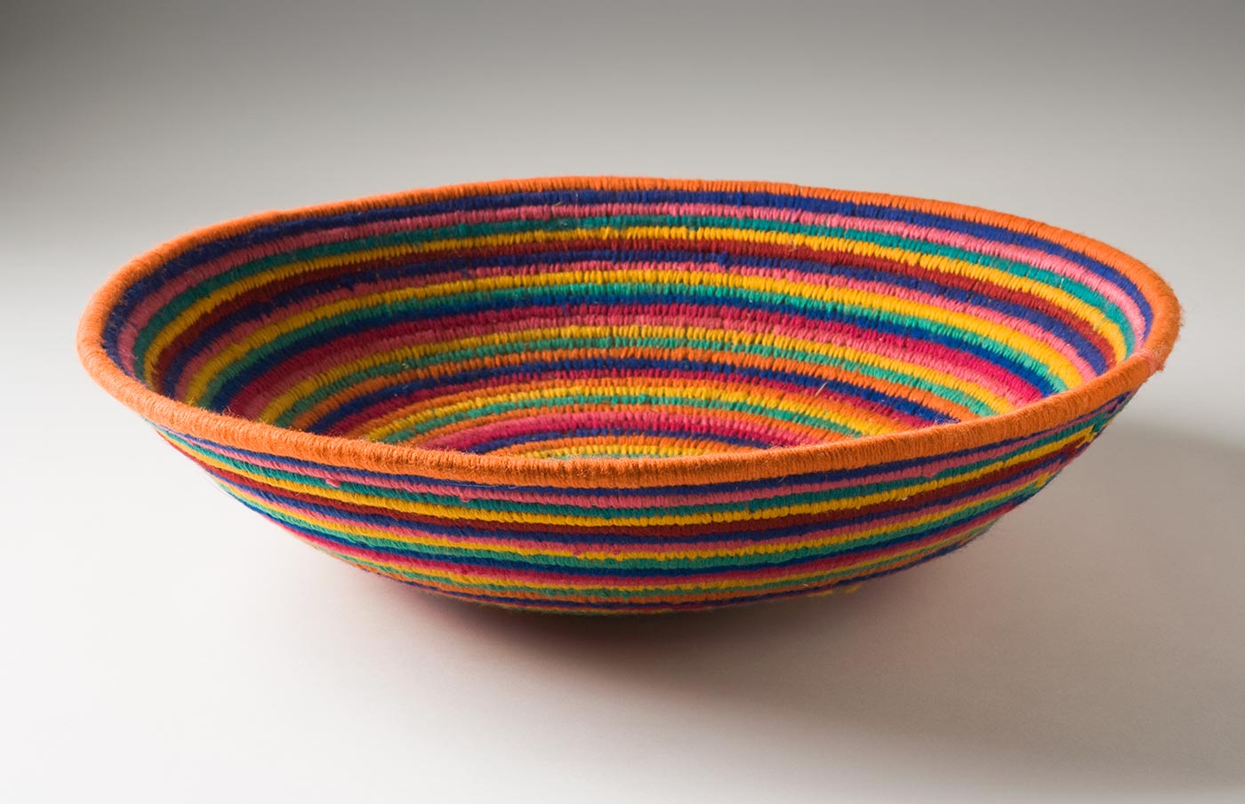 A multicoloured circular coiled yarn and plant fibre bowl-shaped basket. The centre of the basket is in pale pink yarn followed by horizontal stripes of yarn in lavender, pink, green, yellow, orange, blue and dark pink. - click to view larger image