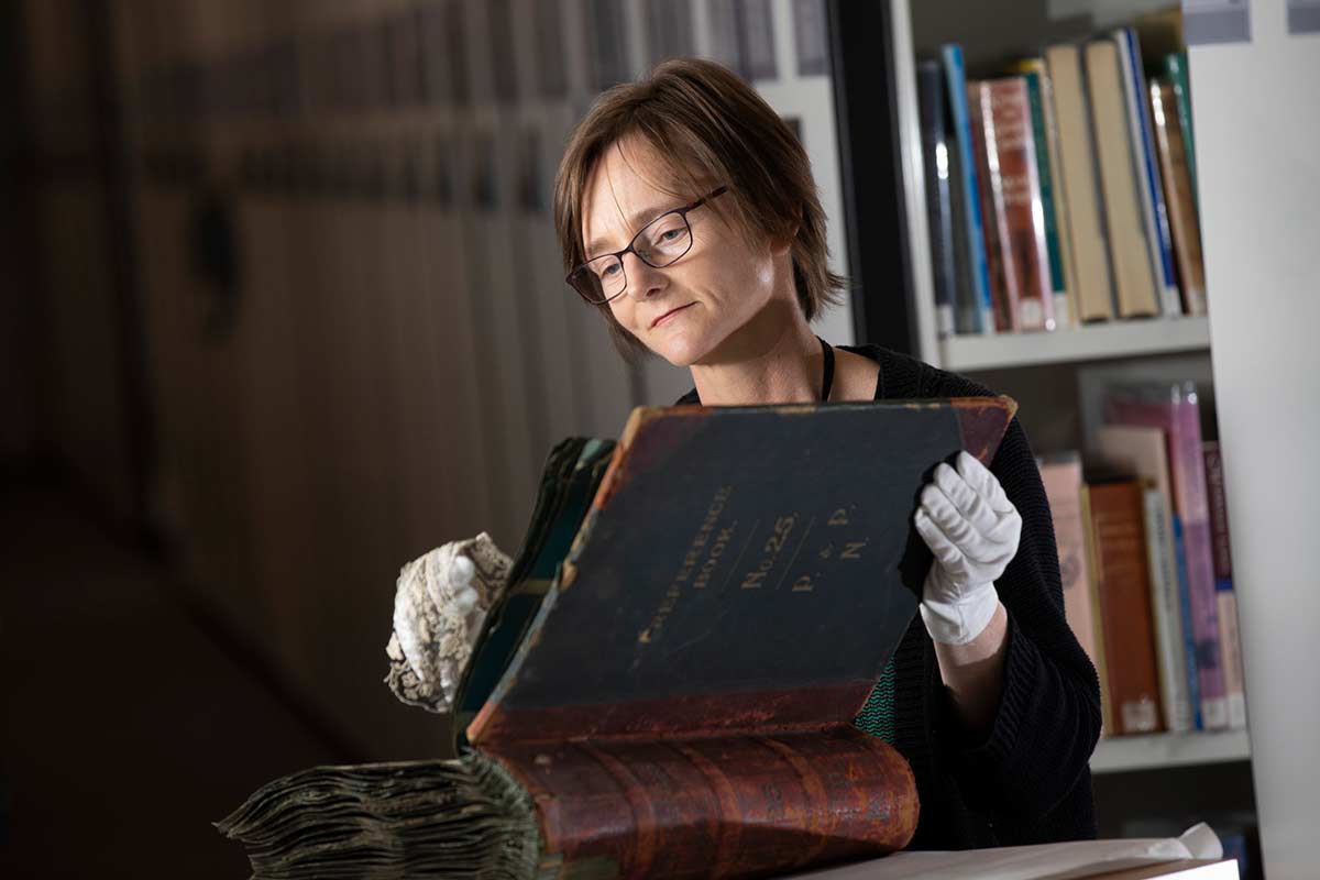 Colour photograph of a woman wearing conservation gloves and studying lace samples from a large leather-bound book. - click to view larger image