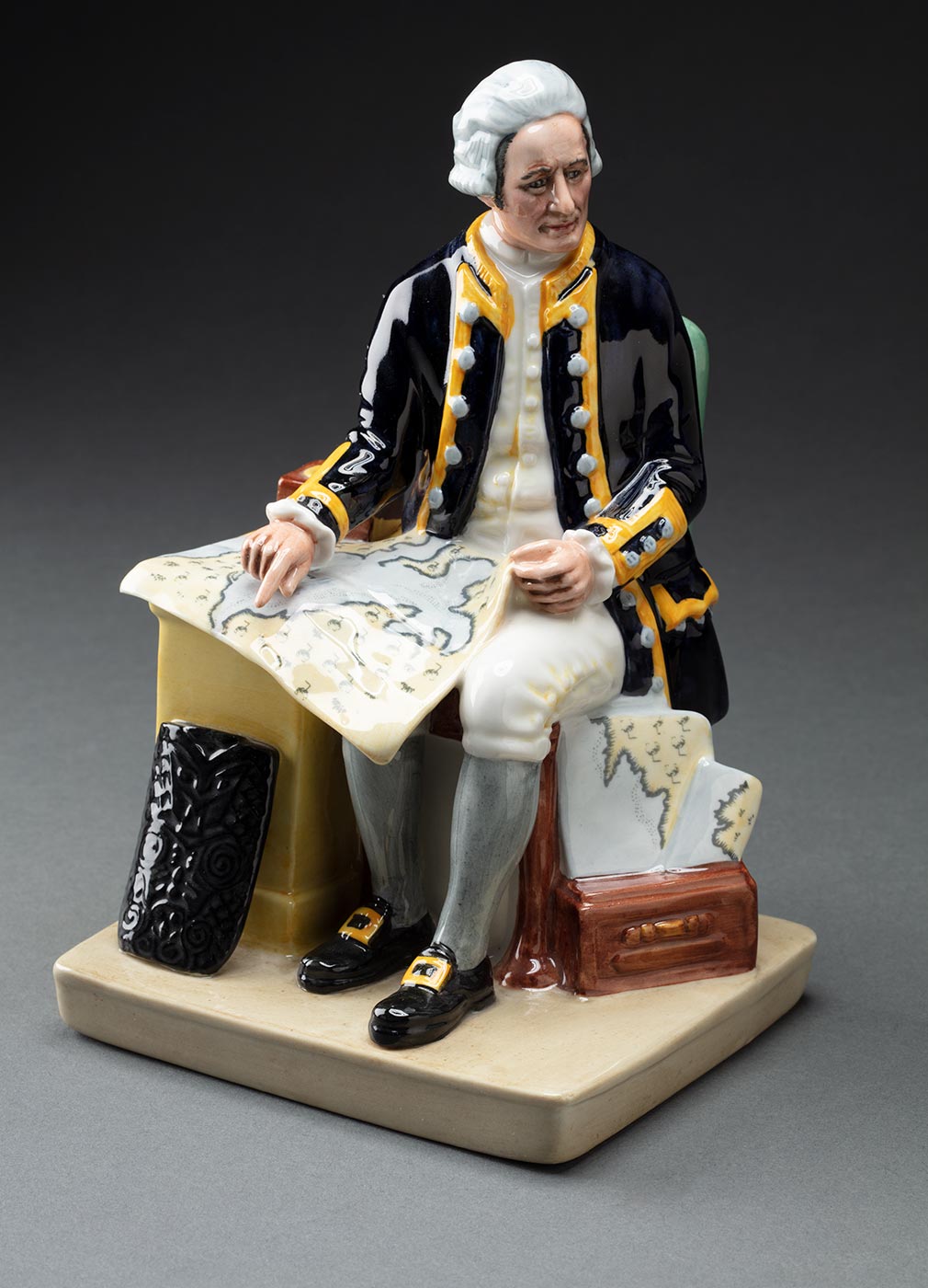 Figurine depicting Captain Cook sitting in a chair. A map is on his lap and he is looking off to the right. A table sits to the left with a compass and a tribal shield. The figurine has a square base, the underside of which is stamped 'Made In England/ Royal Doulton / England/ Captain Cook/ HN 2889/ Royal Doulton/ Tableware Ltd 1979'. - click to view larger image