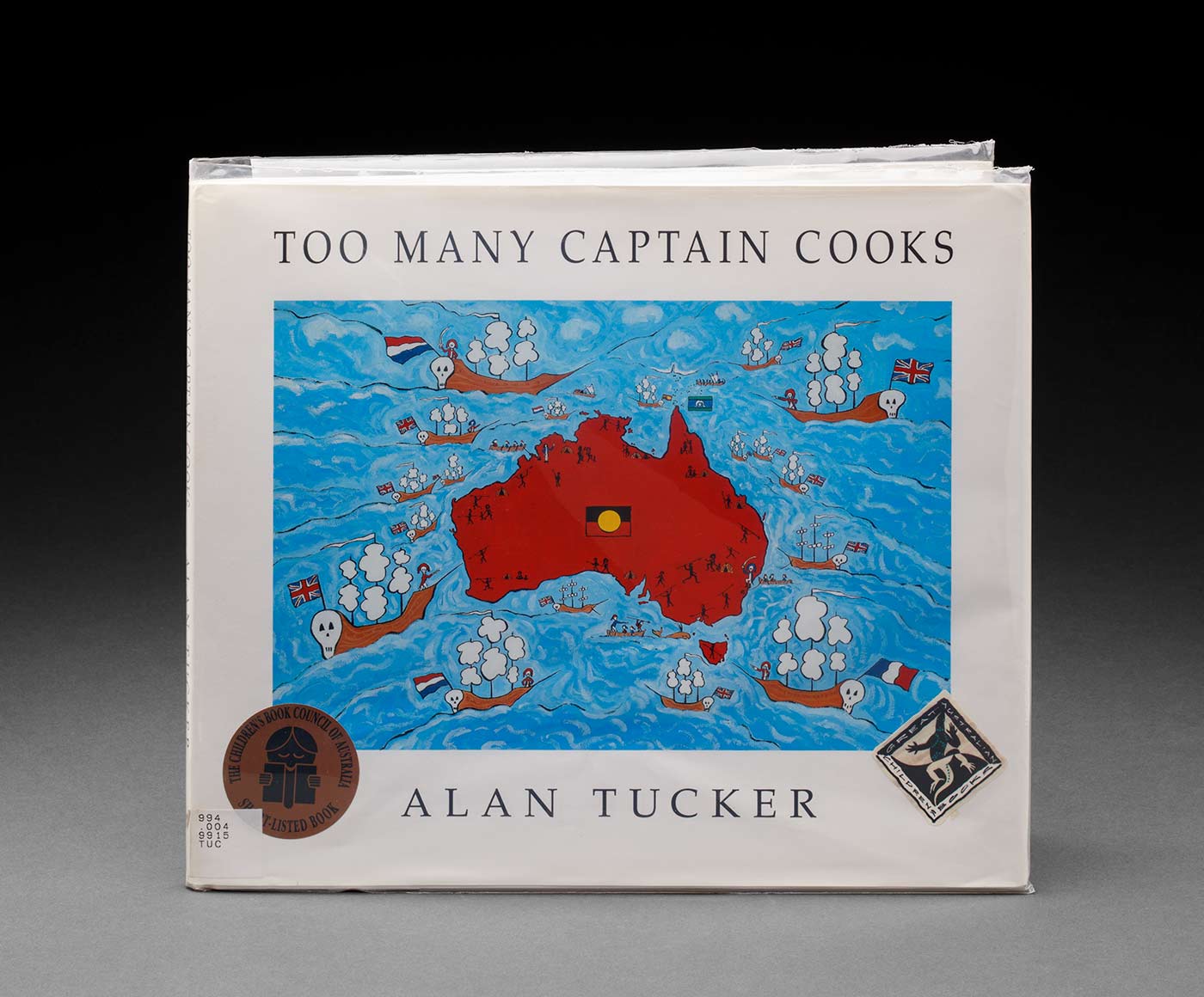 A hard cover children's book titled 'TOO MANY CAPTAIN COOKS' by 'ALAN TUCKER'. The cover features a map of Australia in red surrounded by blue water with a white boarder. There are boats with skulls at one end on the water around Australia and figures around the edge of Australia holding spears. There is an Aboriginal flag in the centre of Australia. - click to view larger image