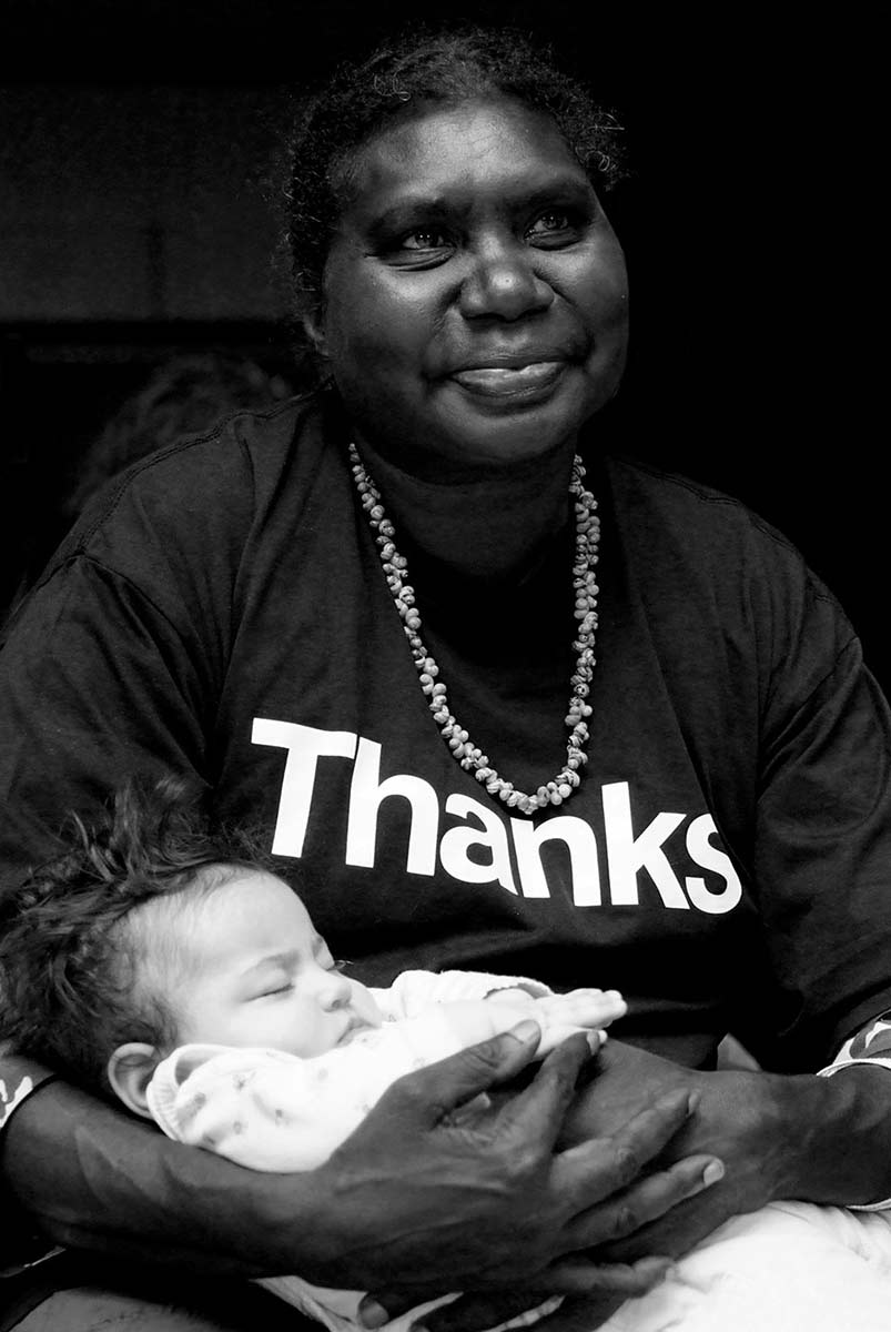 Black and white photograph of a smiling woman holding a sleeping baby. The woman is wearing a T-shirt printed with the text: ‘THANKS’ and a shell necklace around her neck. - click to view larger image