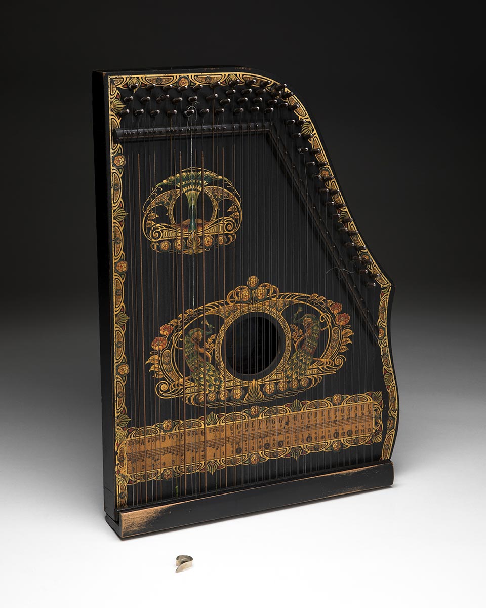 An auto-harp made from wood and metal with a painted exterior. The wooden sound box is designed in a stylised shape with a circular sound hole in the centre. There is a series of 60 strings fixed to the top of the sound box. There is a painterly decoration featuring peacocks, floral designs and a musical score, in gold, brown, green and red hues. The main exterior is painted black. The paint is worn in areas from use. - click to view larger image