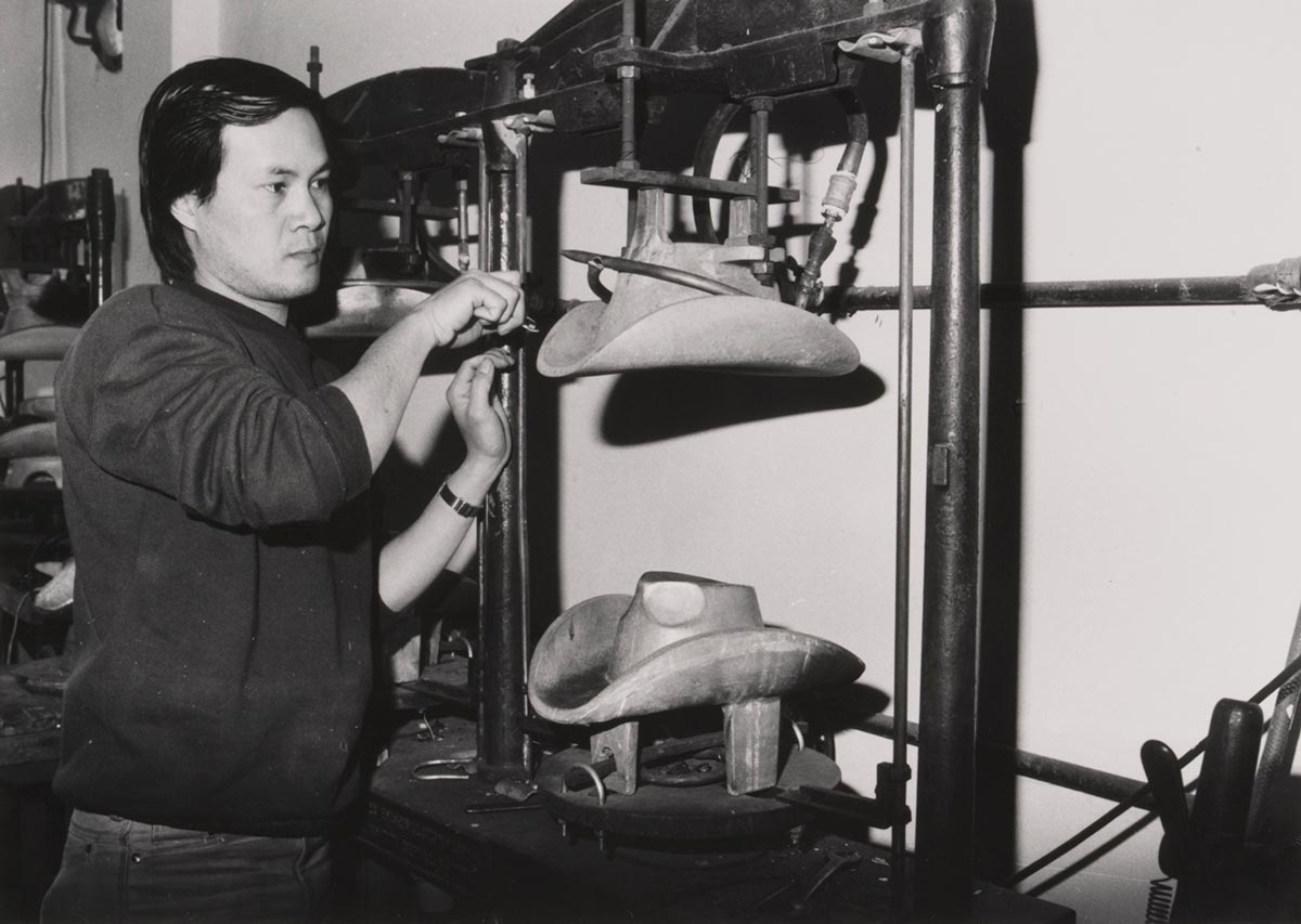 A black and white photograph of a man using a hat blocking machine. The mould attached to the machine is for a cowboy hat. - click to view larger image