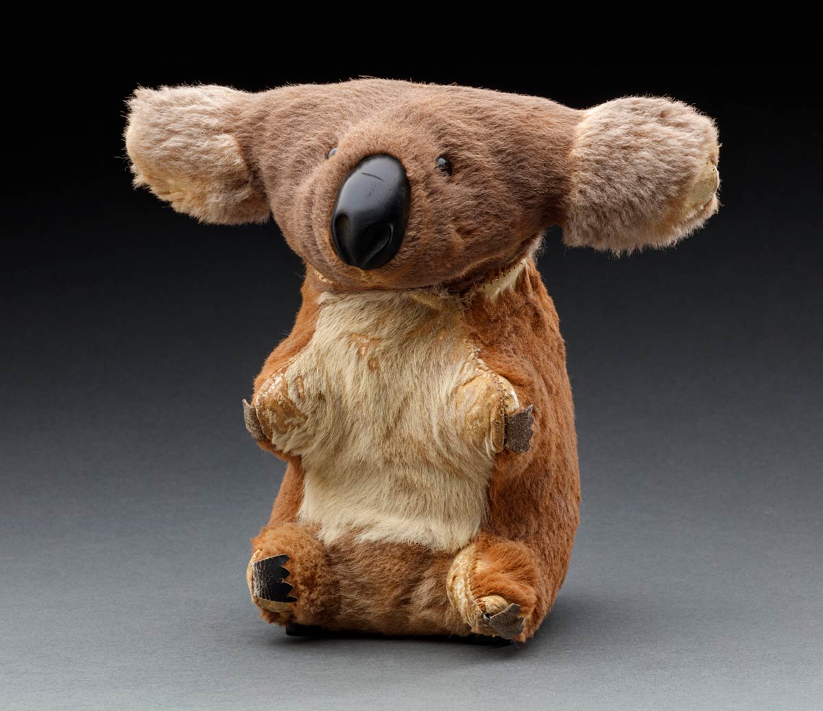 A toy koala made of grey and brown kangaroo fur. It has small black plastic eyes, and black leather for the nose and claws. There are a number of areas across the toy where the fur is missing. - click to view larger image