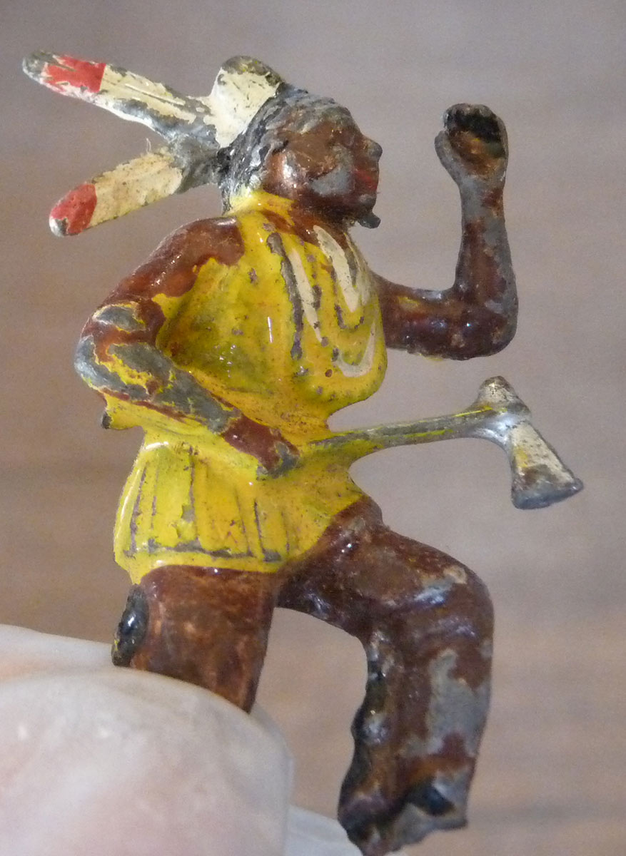 A painted ceramic figurine of a man in Native American attire, holding his left fist up and holding an axe in the other. - click to view larger image