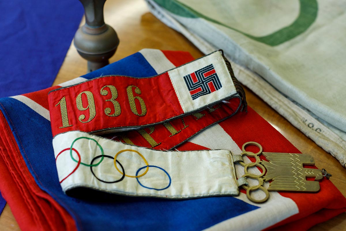 A fabric belt with a print of the Olympic rings and the text: 1936 with the Nazi symbol. There is a gold buckle at one end featuring an eagle perched on the Olympics rings. - click to view larger image