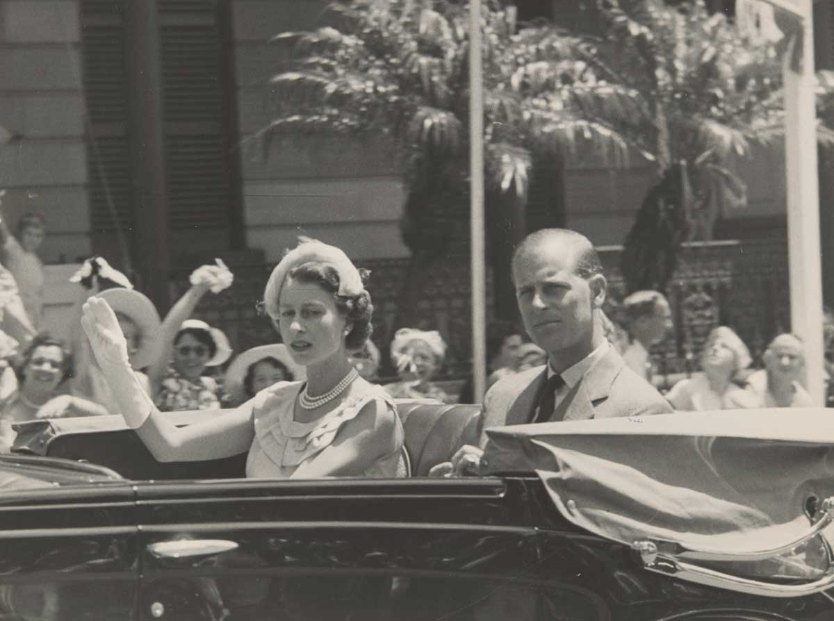 A black and white photograph of Queen Elizabeth II and His Royal Highness The Duke of Edinburgh, in an open car, waiving to the crowd on the street.