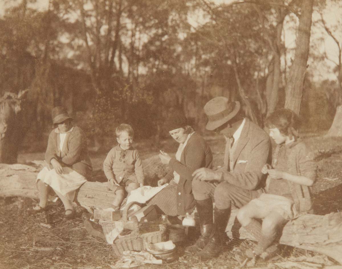 Sepia photograph of three adults and two children sitting on a log, a picnic basket at their feet. - click to view larger image