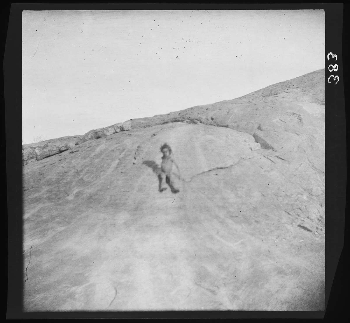 A small First Nations child tobogganing down a bare rocky slope on a seat of rushes, Kurrekapinnya Soakage, Ayers Range, Northern Territory 1903. The surface of the slope has marks that suggest that the tobogganing was a frequent game. The child is in the middle of the image; behind is more rock leading up to a large slope that rises up from left to right in the image. - click to view larger image
