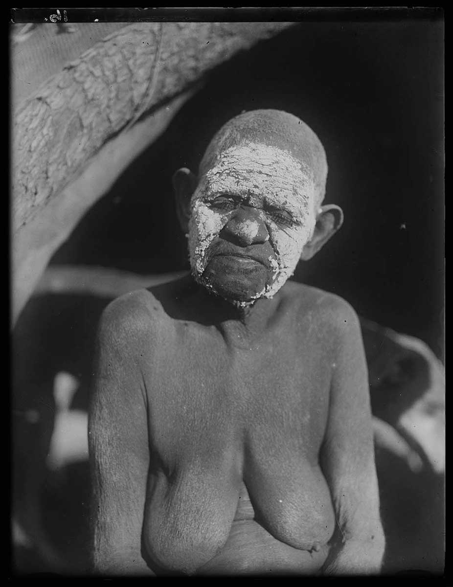 A portrait image of an elderly Aboriginal woman with a painted face and a close-cropped haircut. She sits at the entrance to a hut, with her eyes shut to keep out the sun. The pigment on her face is roughly applied to her forehead, cheeks and the end of her nose. Her hair and scalp are light in colour, as though the pigment has been applied to them. Behind her are deep shadows formed by the interior of the hut. - click to view larger image