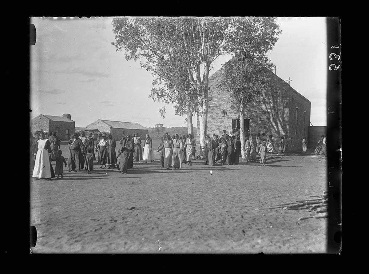 Aboriginal women and children leaving Hermannsburg church, Northern Territory 1920. The church is to the right of the image, behind two trees. The people leave in a large stream, mostly walking right to left across the image. They all wear European cothing. Two other buildings can be seen in the left background. The bare ground in the image foreground is dimpled, possibly from many footsteps and horse hooves. - click to view larger image