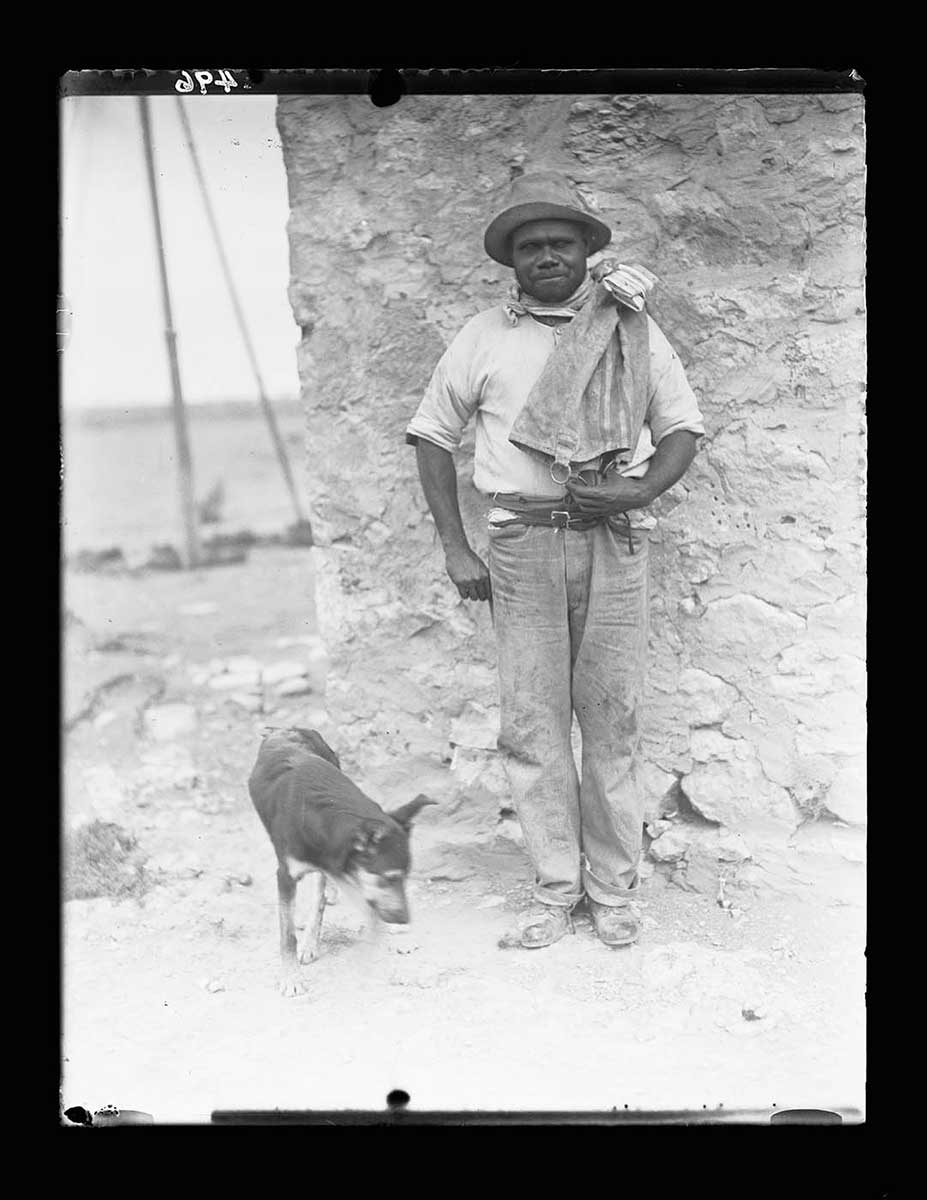 An Aboriginal man standing in front of a building wall, Nullarbor Plain, South Australia 1920. He wears trousers, an undershirt and a hat. He has what appears to be a mailbag over his left shoulder. A dog stands at the man's right side, looking at the ground. The wall is made from earthen materials. To the left of the image the horizon can be seen. - click to view larger image