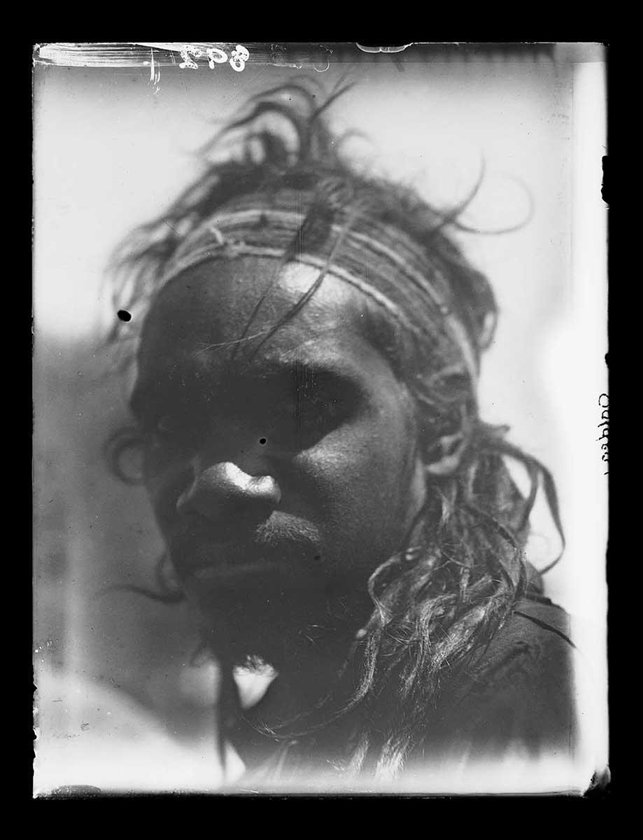 Portrait of a young Aboriginal man, South Australia 1920. He wears a headband high on his forehead. His long hair hangs down on his neck and shoulders. He faces the left side of the image with his head turned toward the camera. He appears to wear a coat or heavy shirt. His face is in deep shadow, however his eyes can still be seen looking at the camera. The background is out of focus, adding emphasis to the young Aboriginal man. - click to view larger image
