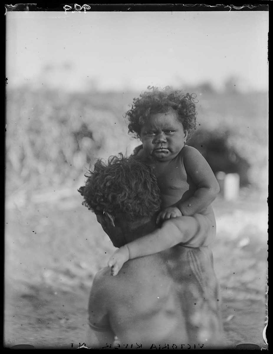 Aboriginal man carrying a Aboriginal child on his shoulder, Victoria River, Northern Territory 1922. The man has his back to the camera and is only visible to his lower back. The child is perched on his right shoulder, with its left leg around the back of the man's neck. The child looks directly at the camera, its cherubic face looking a little stern. The background is out of focus. Some blurred objects in the right background may be a hut with a box or case in front of it. - click to view larger image