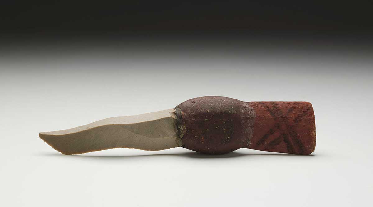A stone flake-bladed knife photographed against a neutral grey studio background. The stone flake blade has a triangular cross-section and a wavy cutting edge. A football-like section of natural resin with flattened sides connects the blade to a short wooden handle. The handle has on it a pattern forming a single 'x' design. - click to view larger image