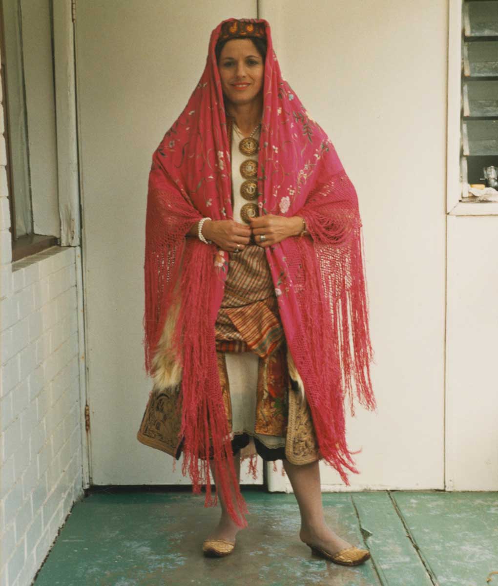 Portrait photograph of a woman wearing a traditional Greek garment with many layers. She wears a knee-length off-white chemise over a pair of knee-length pantaloons. Over the chemise is a crimson knee-length tunic embellished with gold thread and colourful silk embroidered floral motifs.  Wrapped around her waist and hips is a woven sash with fine checked pattern. Worn over the sash and tunic is a dark coloured three-quarter length overcoat with cream-coloured fur trim down the front opening and hems of the sleeves. The woman wears a yellow, red and blue beanie-shaped hat, and a bright pink silk shawl with long fringed edges is draped over her head and shoulders. The woman wears a pair of embroidered gold slippers with a small heel. - click to view larger image