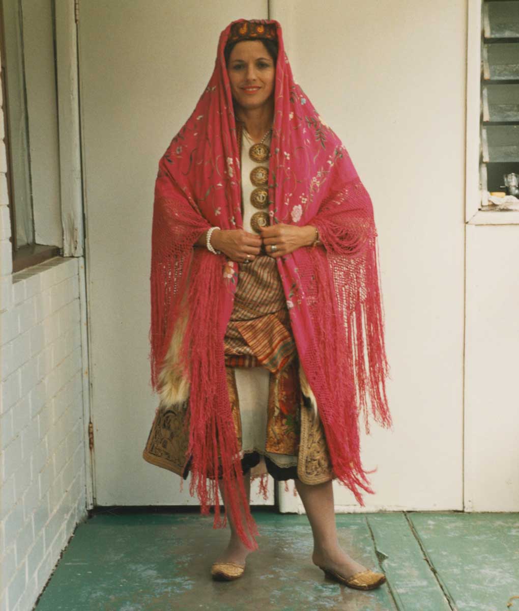 Portrait photograph of a woman wearing a traditional Greek garment with many layers. She wears a knee-length off-white chemise over a pair of knee-length pantaloons. Over the chemise is a crimson knee-length tunic embellished with gold thread and colourful silk embroidered floral motifs. Wrapped around her waist and hips is a woven sash with fine checked pattern. Worn over the sash and tunic is a dark coloured three-quarter length overcoat with cream-coloured fur trim down the front opening and hems of the sleeves. The woman wears a yellow, red and blue beanie-shaped hat, and a bright pink silk shawl with long fringed edges is draped over her head and shoulders. The woman wears a pair of embroidered gold slippers with a small heel. - click to view larger image