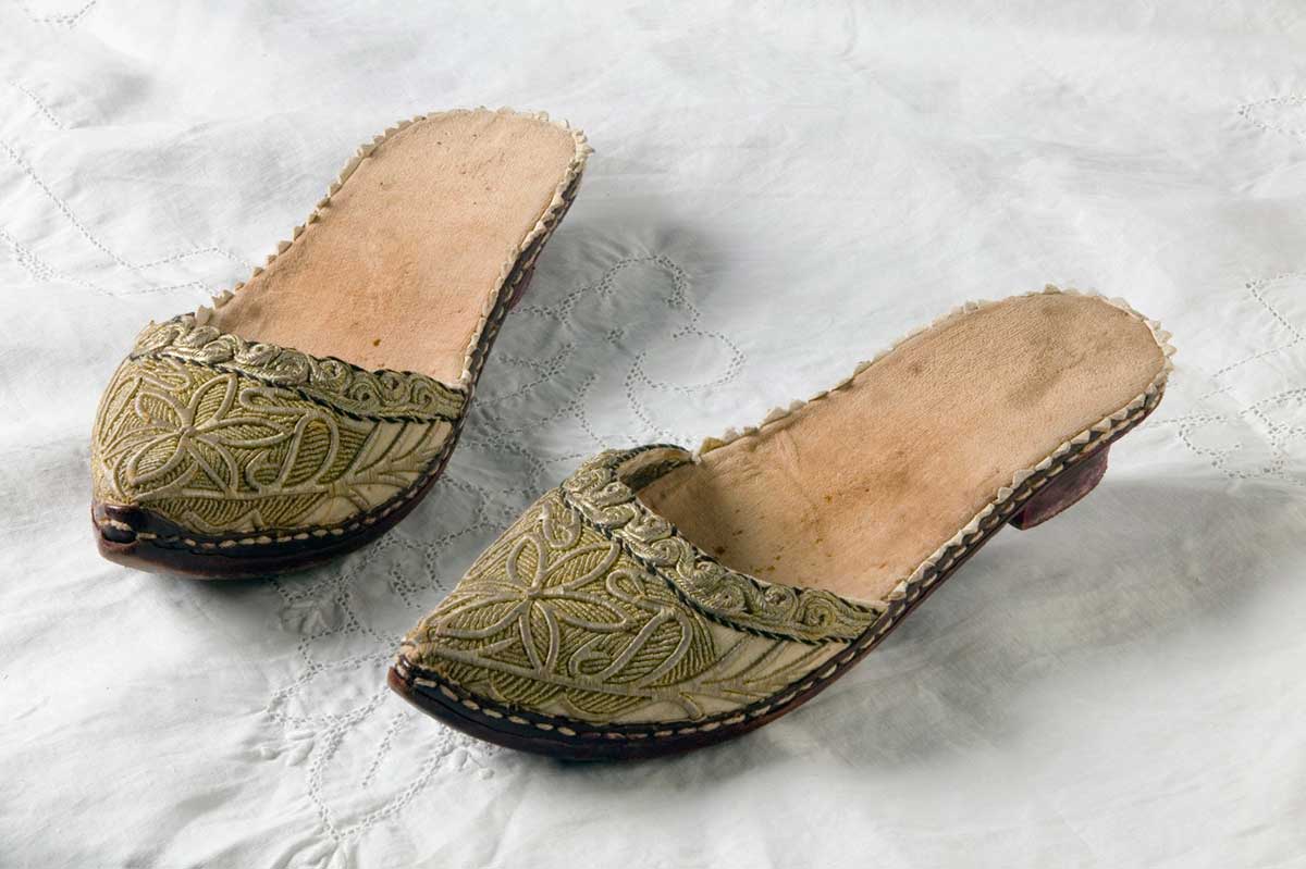 Photograph of a pair of Greek backless slippers with a small heel. The slippers are covered in cream-coloured velvet and heavily embroidered with gold thread in a daisy motif with a swirling border. The slippers have a slightly pointed toe. They are photographed on a piece of white cloth. - click to view larger image