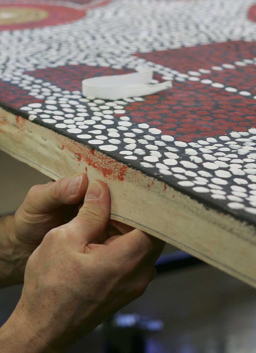 A conservator uses his hands to pull edge of canvas over timber frame. - click to view larger image