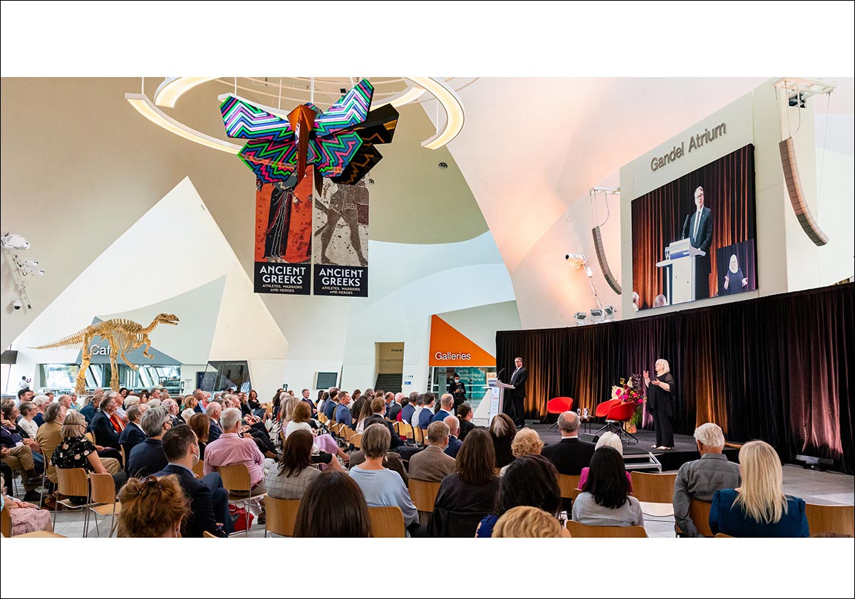 An audience is gathered in the Gandel Atrium of the National Museum of Australia, watching a man presenting on stage. - click to view larger image