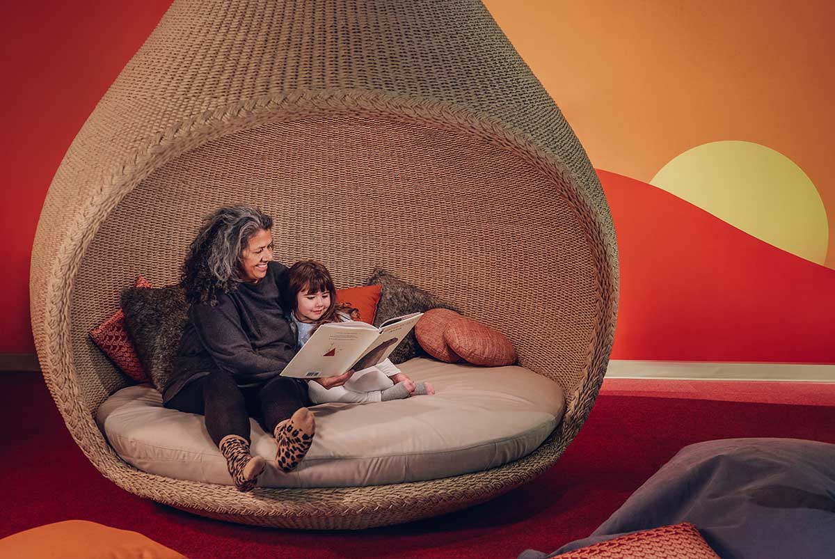 A woman and a girl sit reading in a large woven vessel shaped like a pod. - click to view larger image