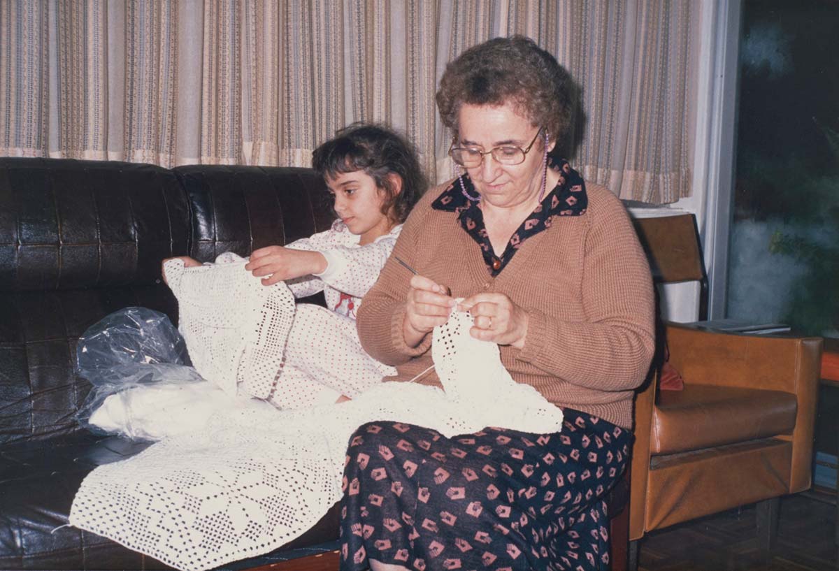 Coloured photograph of Effie Kyprios sitting on a black leather couch, crocheting a white bedspread. Effie has short, grey, curly hair, and is wearing a purple dress with a pink square print, and a brown knitted cardigan. She wears glasses and is looking down at her hands, which are holding the bedspread and her crochet hook. Effie’s young granddaughter is sitting on the couch behind her, holding a length of the bedspread in both her hands, looking at the design. She has dark hair and wears a white polka dot top. - click to view larger image