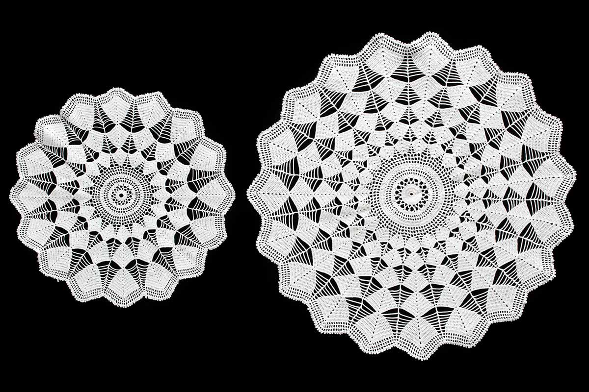 Set of two round white crocheted table settings on a black background. One piece is smaller than the other. The table settings have a detailed web-like design in concentric circles and have zigzag edges.  - click to view larger image
