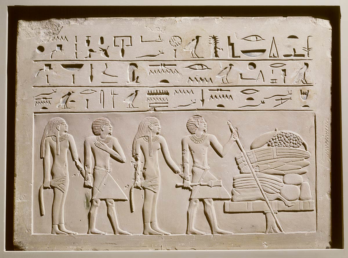 Rectangular stone with three rows of hieroglyphics carved at the top and four figures, in relief, beside a table laden with offerings as the main image.