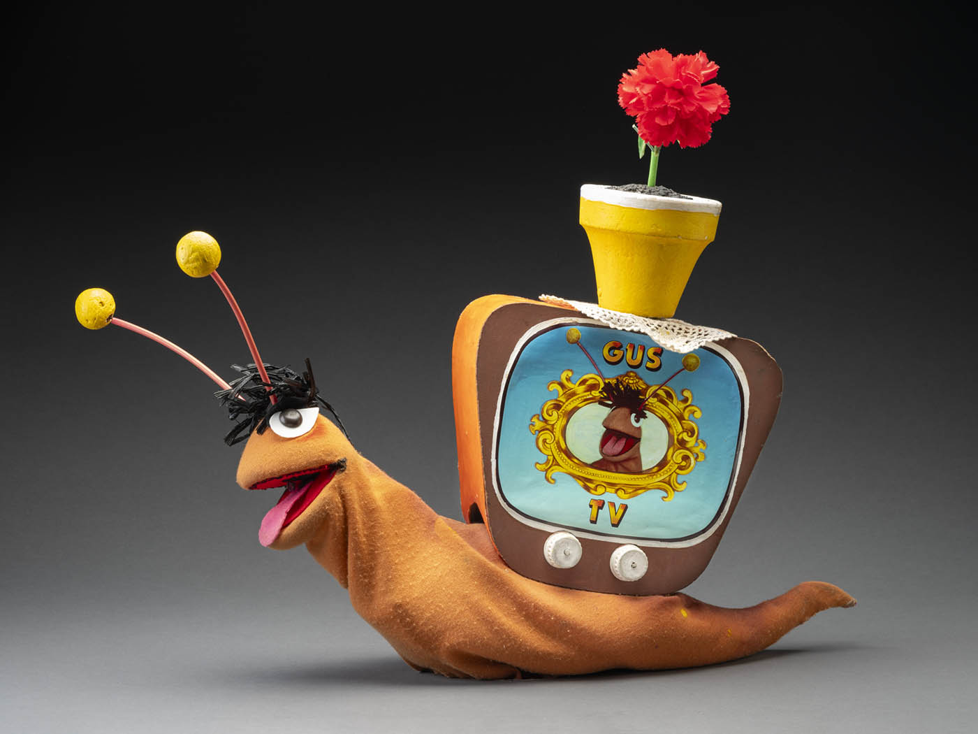 A puppet of a snail named Gus. Gus has an orange body, a tuft of black hair, orange and yellow antennas, brown eyes and a pink felt tongue. Attached to its back is a television box with two knobs, a screen featuring an image of Gus’s head surrounded by a gold frame, and printed text that reads ‘GUS/TV’. Attached to the top of the television is a yellow flowerpot with a red flower, sitting atop a doily. 