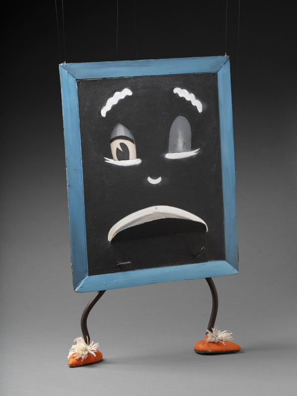 A blackboard puppet with two legs at the bottom and strings attached at the top. The board features a face with a blue frame and two protruding pegs. There is a string attached to the mouth which opens outward. Attached to the legs are orange painted shoes with white pom-poms and green felt on the bottom.