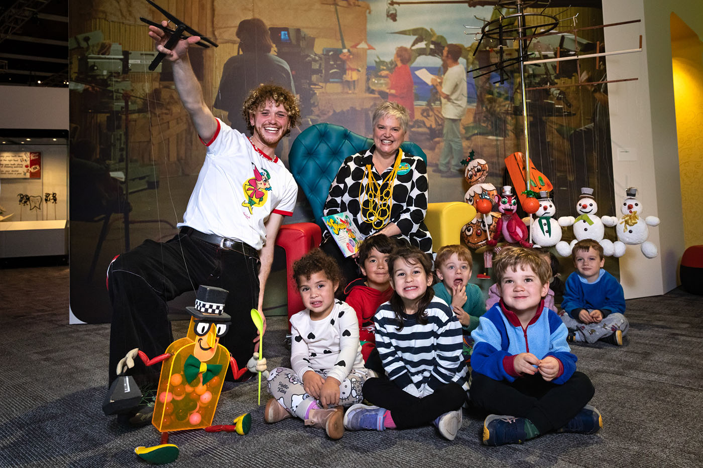 A man holding a puppet shaped like a bottle kneels alongside a group of small children and a woman, sitting on a chair behind.
