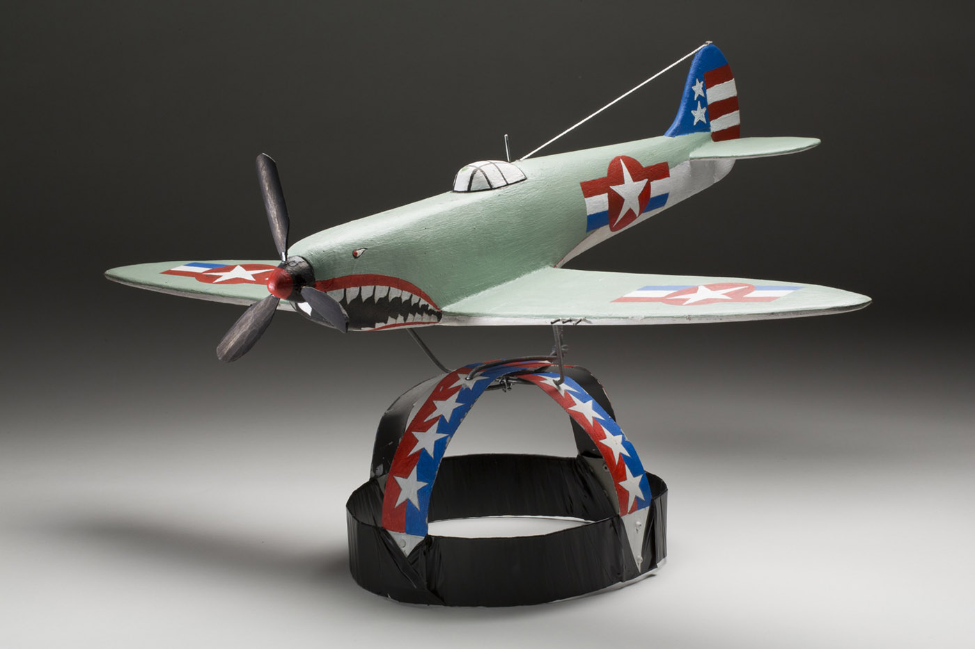 A headdress with a American fighter plane, circa World War 2 as the feature. The headpiece is painted with white stars against a red and blue striped background. On top of the headpiece is a painted wooden model of an American plane. The plane is painted green with star and stripe markings in blue, red, and white, and the nose features a propeller and a face design with an open mouth showing sharp teeth. - click to view larger image