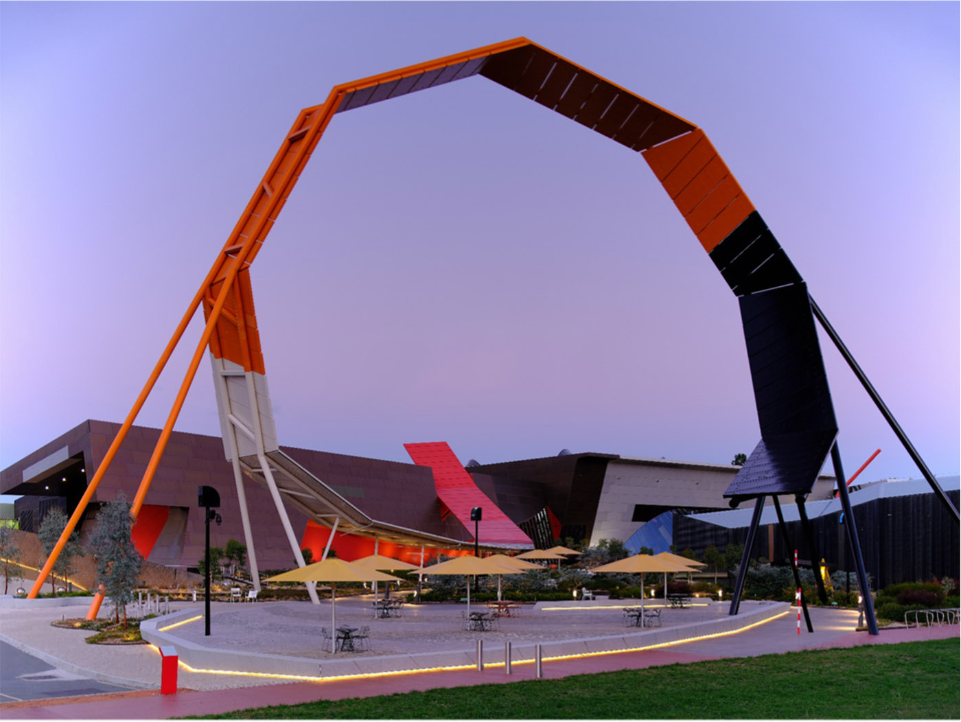 A photograph of the Fore court at the Museum. The Museums colourful loop is above a paved area. The paved area has umbrellas and tables.