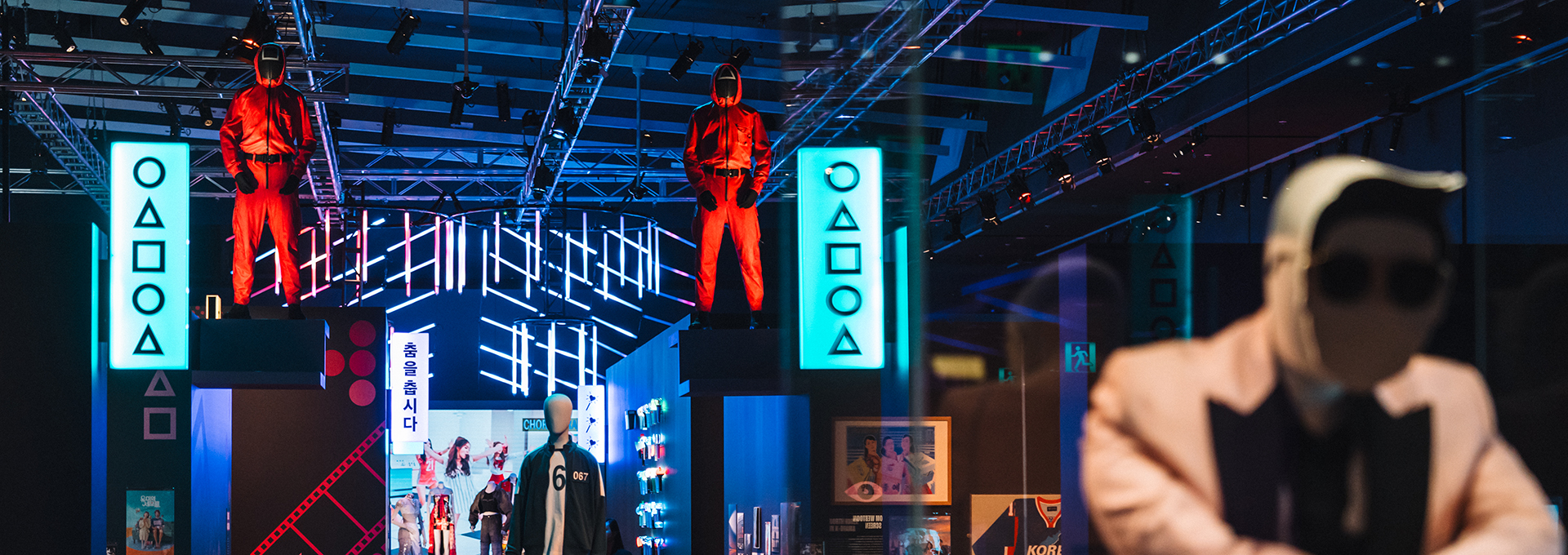 A photograph of the Hallyu exhibition, focusing on the Squid game tracksuits on mannequins. The mannequins are amongst the colourful exhibition space which has lots of lights and screens.