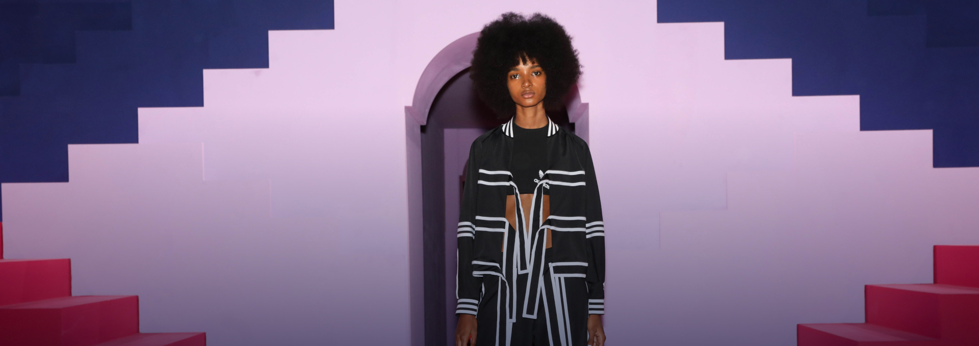 Photo of a model wearing a black and white Korean influenced Adidas track suit, standing in front of a purple arched doorway with pink steps and navy-blue background.