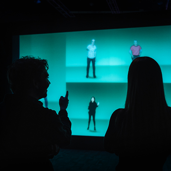 Two people stand looking at a screen with images. Their back are to the camera and they are in the shadows. On the screen are people dancing on a green background.
